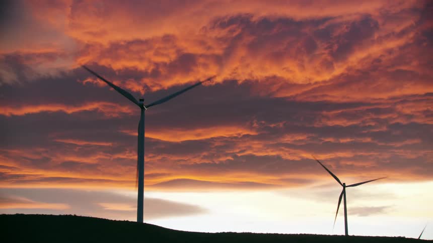 Two silhouetted wind turbines rotate on a ridge with vibrant orange purple and pink sunset clouds in the background as clean electricty is produced in beautiful conditions
