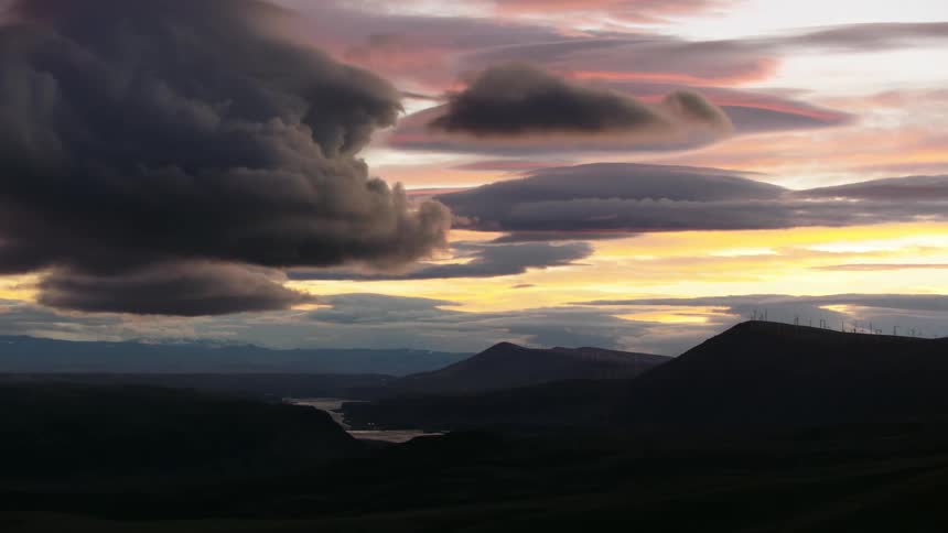 Looking across a vast river valley with outrageous sunset clouds painting the sky in technicolor hues and tones with windmills on a ridge on the right side of the frame