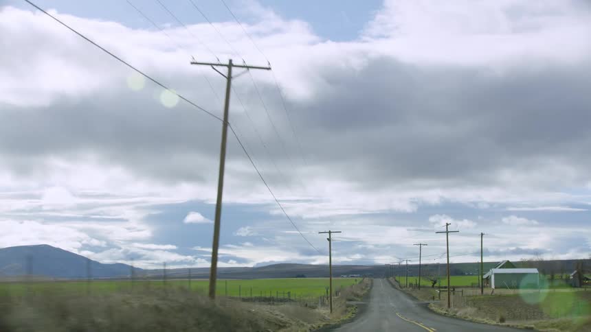 A two lane road runs past a farm house and large open fields in early spring with vast sky and white puffy clouds in the blue sky