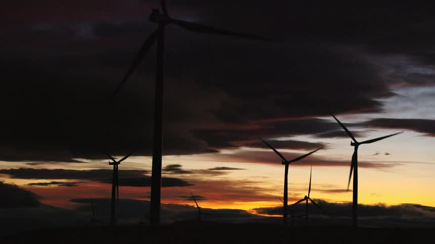 Many wind turbines rotate with purpose silhouetted by an outrageous sunset sky filled with orange, blue and purple light as renewable and clean energy is being generated