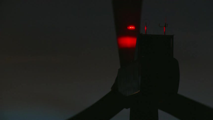 Tight shot of a wind turbine at night with a blinking red light punctuating the camera move past the whirling blades on the windmill