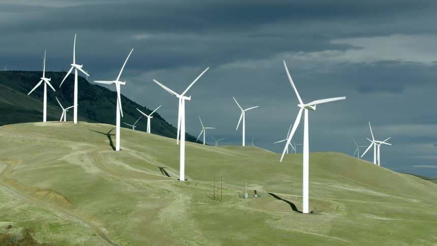 Wind turbines spinning on a wind farm while afternoon sun illuminates the white wind mills with dark high altitude clouds in the background as they create clean electrcity
