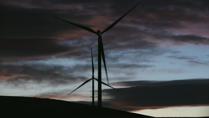 Two wind turbines rotate with vigor and purpose silhouetted in front of a beautiful late evening sky filled with orange and various tones of blue sky