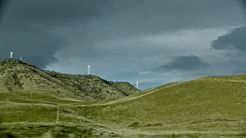Wind mills generate electricity high atop of mountain ridge as seen from the view looking out the window of a car driving on the highway with dark storm clouds in the sky