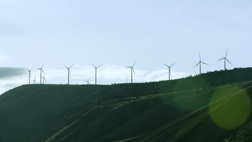 Slow zoom out reveal of over ten wind turbines rotating on top of a large ridge on a big wind farm generating a lot of clean and renewable energy