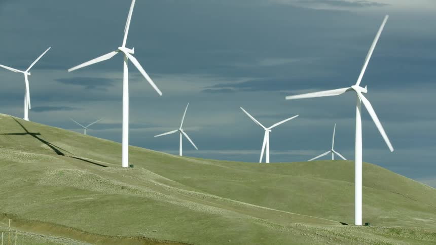 White wind turbines rotate on a large hill at a major wind farm generating clean green renewable electricity for electric utility customers and there are dark storm clouds in the background