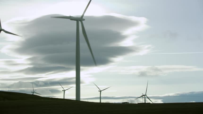 Lenticular clouds form as wind turbines rotate with vigor and speed as the camera passes by as seen from the passenger window of a moving car