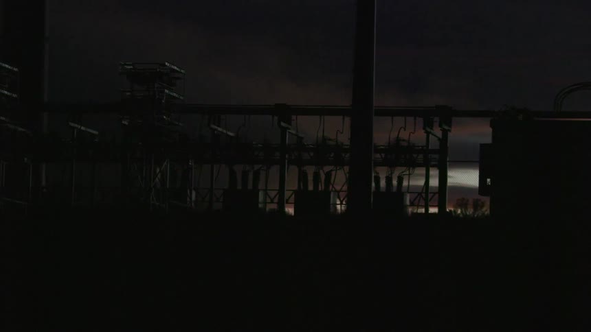 The camera pans left following a power sub station after the sun has set and wind turbines can be seen moving past the background