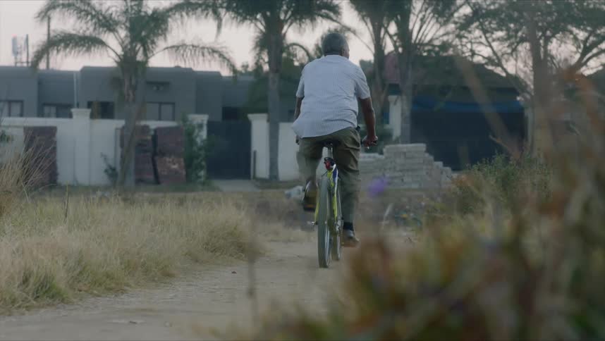 A mature man rides a bicycle at dusk along a dirt path toward small buildings