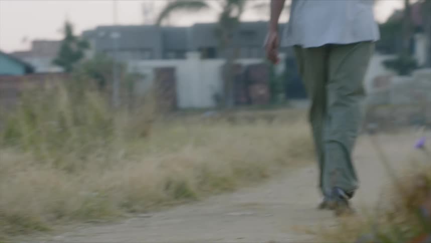 A black man walks down a dirt path near buildings