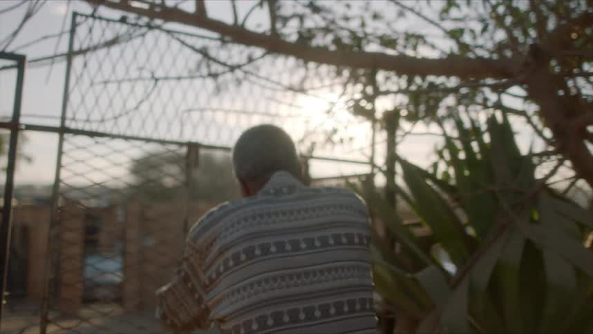 A black man walks along a dirt road and passes through a gate towards a shop in a rural area