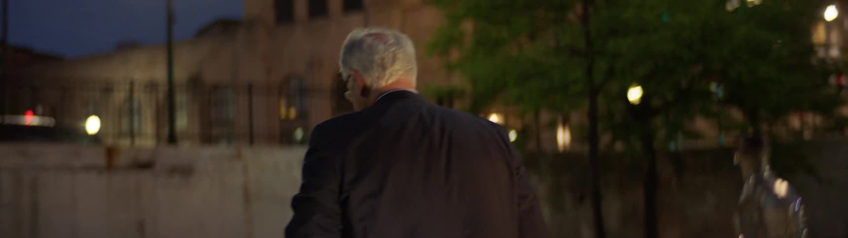 An elderly white man in a suit drinks liquor in the middle of a street in a city at night