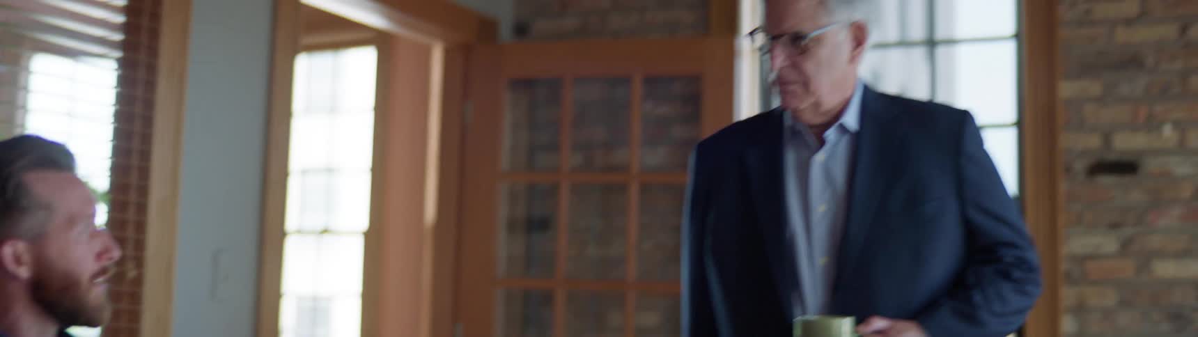 A boss stands at the head of a table looking angry as he listens to an employee talk