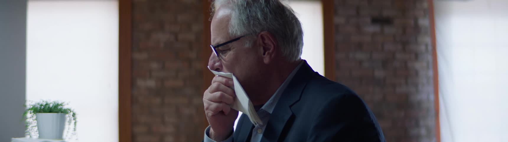 An elderly white man in a suit wipes his mouth with a napkin in an office break room