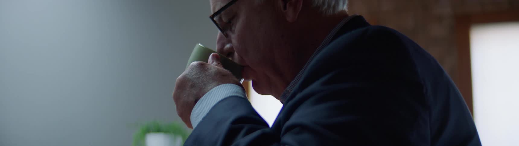 Male standing in break room in front of a table with a coffee maker is sipping on a cup of coffee