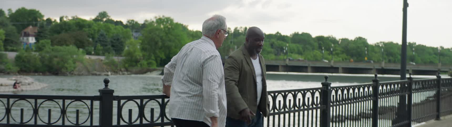 Two elderly men stand and look out at the water while talking and then walk away