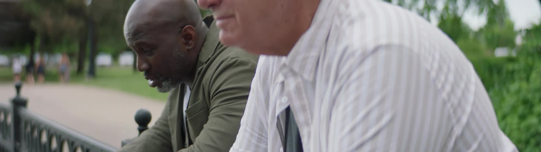 Two male friends lean against a railing along the walkway in the park as they have a conversation together and start to laugh
