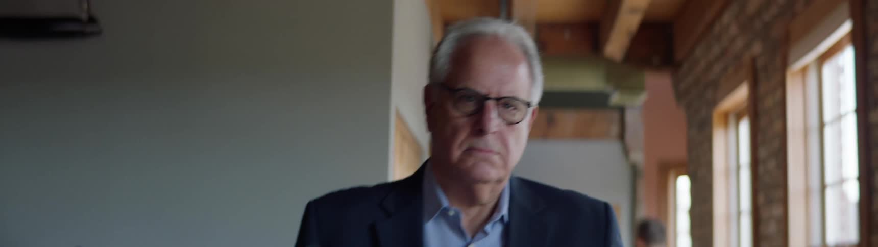 A frowning old man walking through the halls of a building carrying a cardboard box full of desk materials