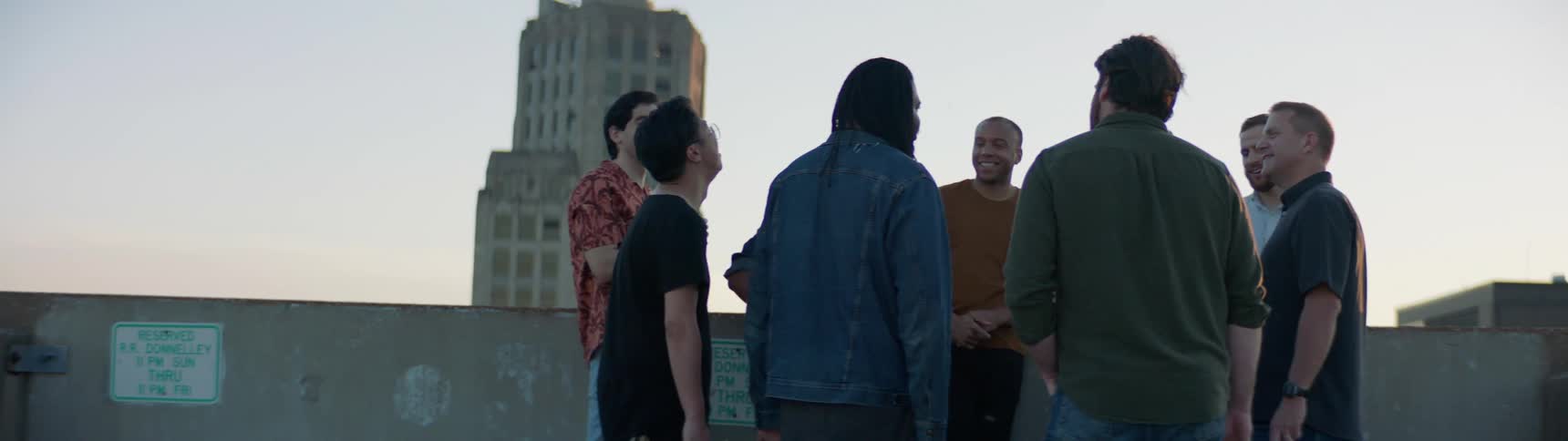 A group of friends laugh as they chat while standing on a building rooftop at dusk