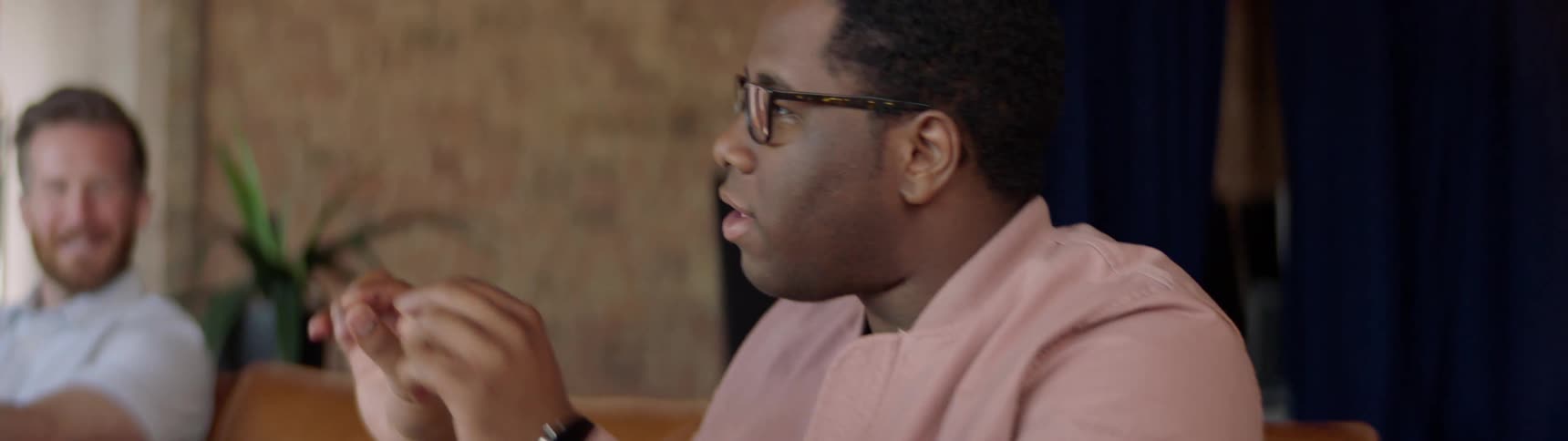 Black man with glasses and a pink coat sits on sofa and talks to his friend by using body language