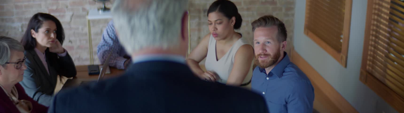 A group of coworkers sit at a table as one man yells at the boss who stands at the head of the table