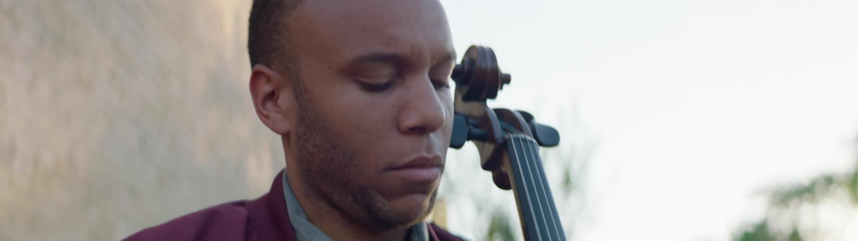 Male sitting outdoors next to a building while playing the cello