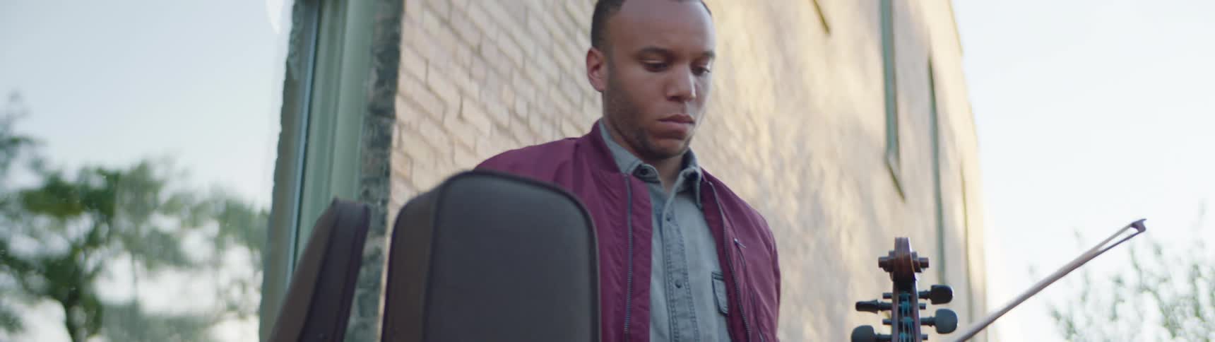 A black man removing a violin and its bow from a case while standing in front of a building