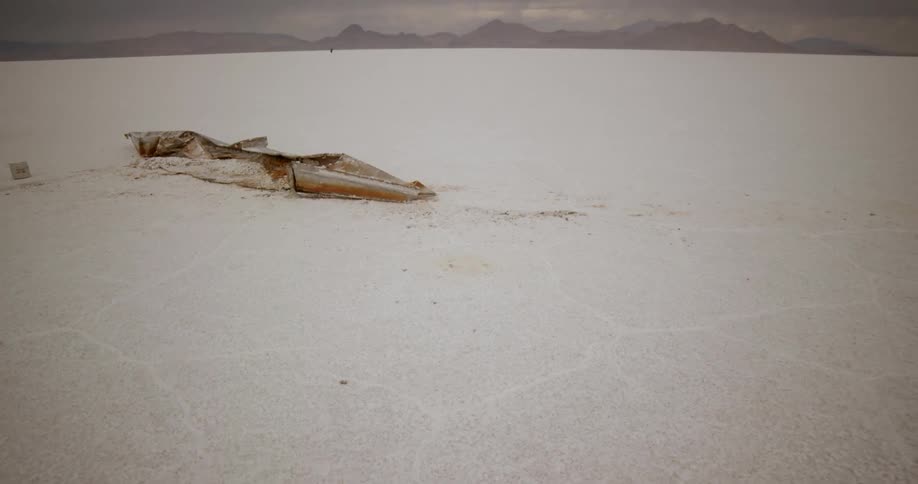 A wrecked boat sits in a desert with a person walking in the far distance