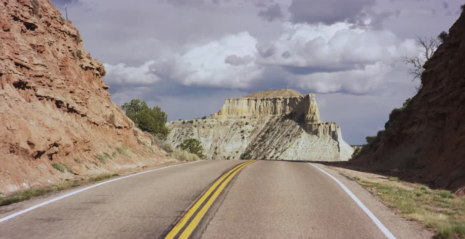 Two lane road passes steep red dirt slopes toward gray mountain under overcast sky