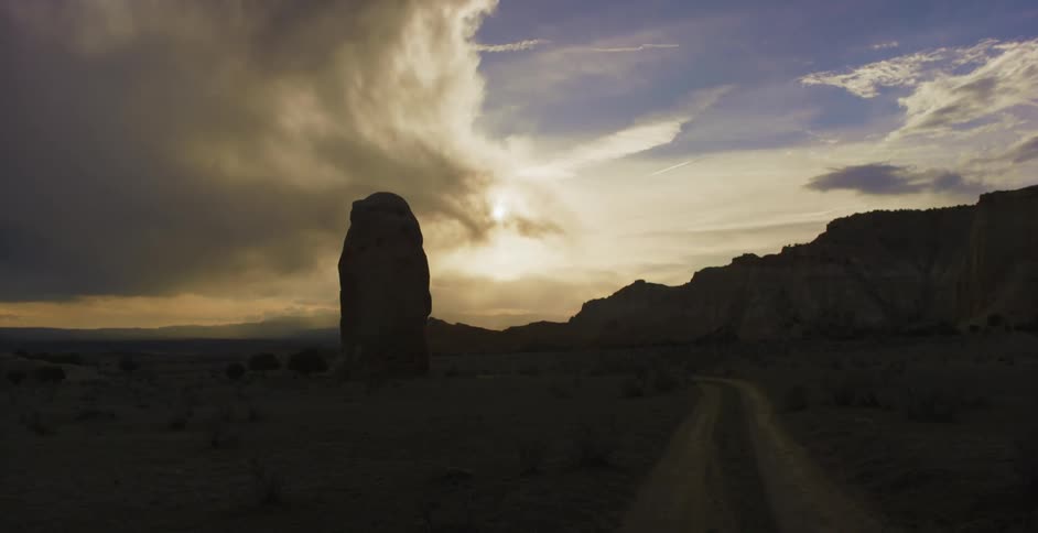A rock formation rises out of a desert near a dirt road and mountains