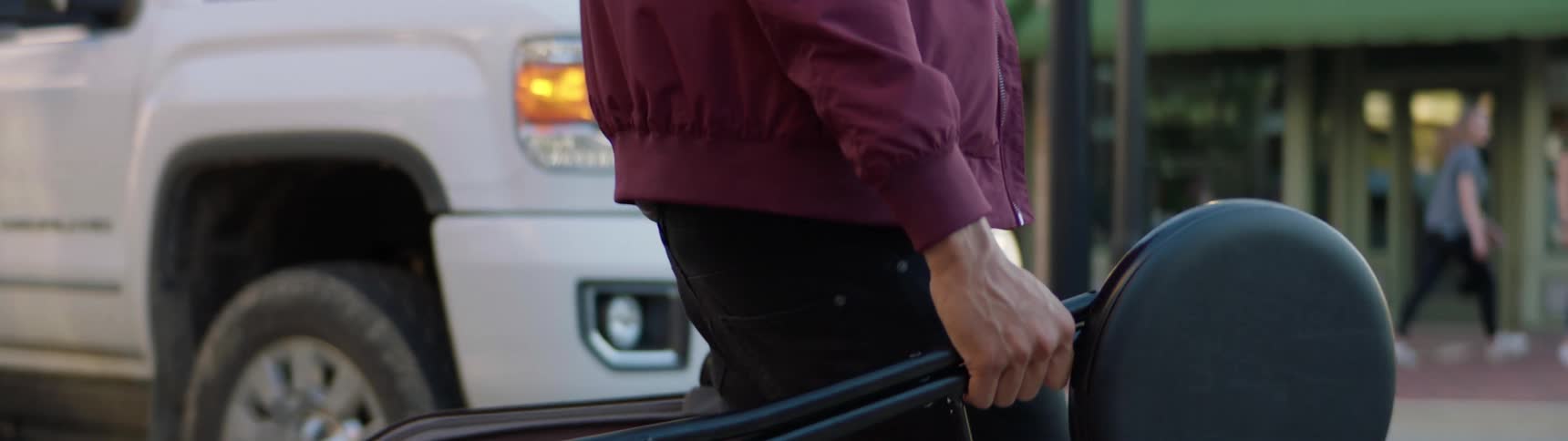 A man carries a musical instrument and stool across a street as people walk along the sidewalk