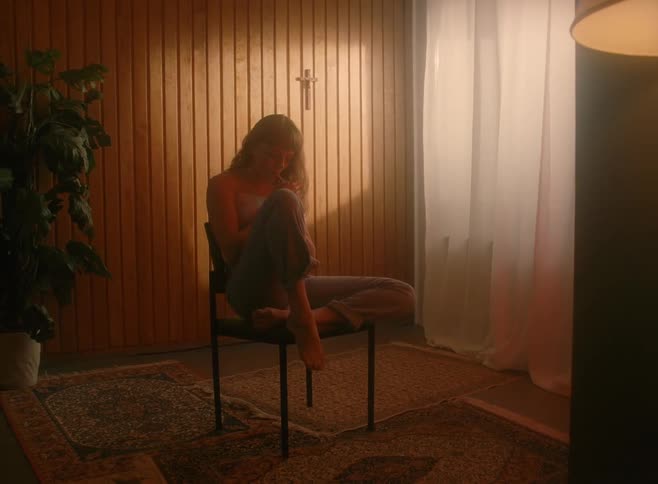 A woman sits in a chair in a dark room smoking a cigarette with a cross hanging on the wall behind her