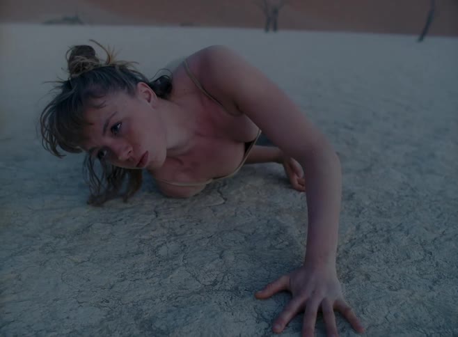 A model writhes on the ground in a desert wearing a nude colored dress