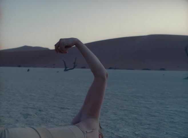 A white woman performs odd dance moves while writhing on the ground in a desert