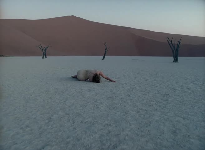 Female dancer lies down on sand in desert with dead trees in the background
