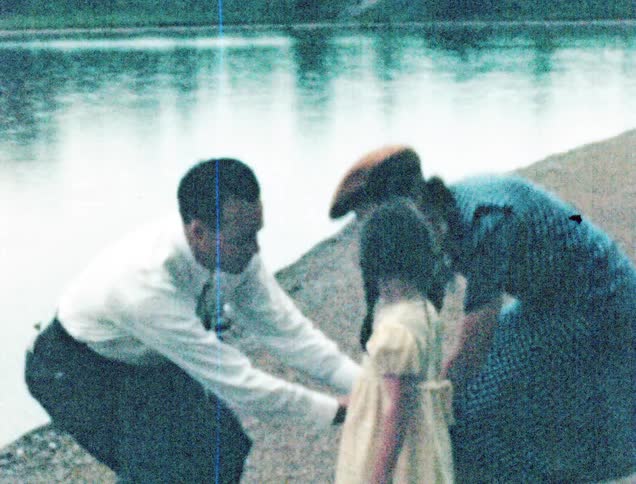 A mother and father put their hands together and bend down for their daughter on the shore of a lake