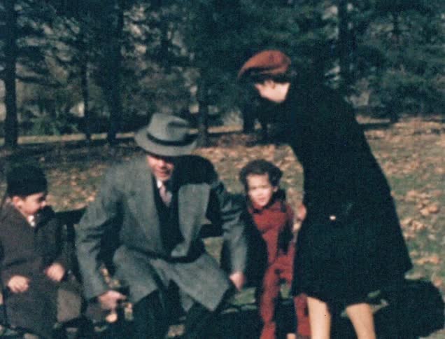 A mother and daughter sit on a bench outside with their three children