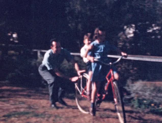 A girl attempting to ride a bicycle while her younger brother sitting behind her and her father pushing the bicycle from behind