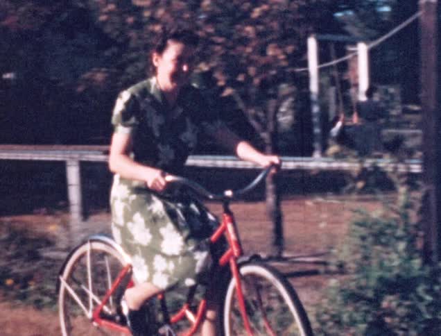 A laughing white woman in a dress rides a red bicycle in a backyard
