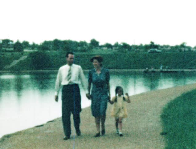 A family of three walk along the sidewalk of an artificial lake in a public park during the day