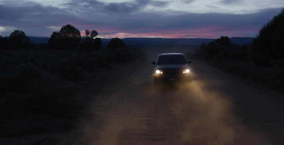 The headlights of a vehicle illuminating clouds of dust as the vehicle travels down a dirt road through the countryside