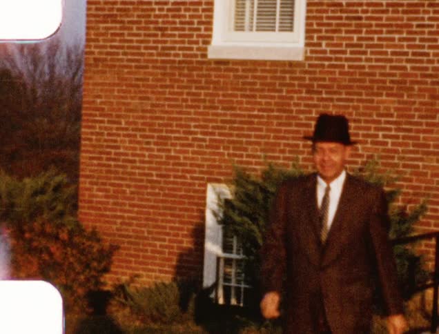 A man with a suit and tie and a hat walking with houses in the background