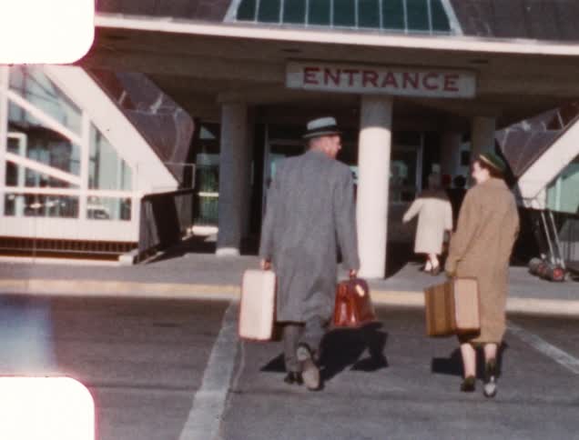 A man waves good bye as him and his wife cross a street with their suitcases