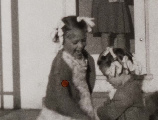 Two little girls play outdoors while a woman watches from a door