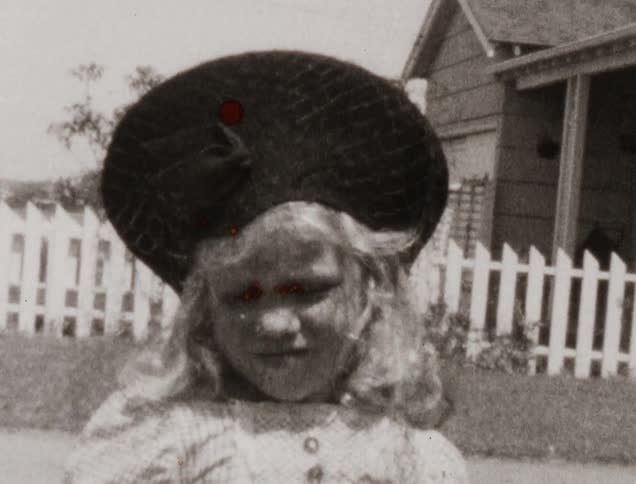 A young girl poses in a dress and large wool hat outside her house