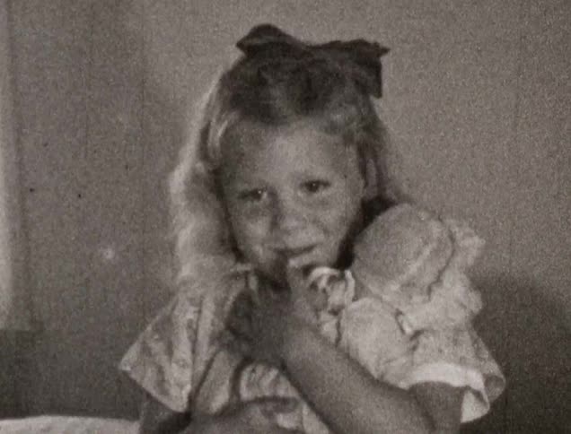 Black and white film of a girl in a dress playing with a doll in a room