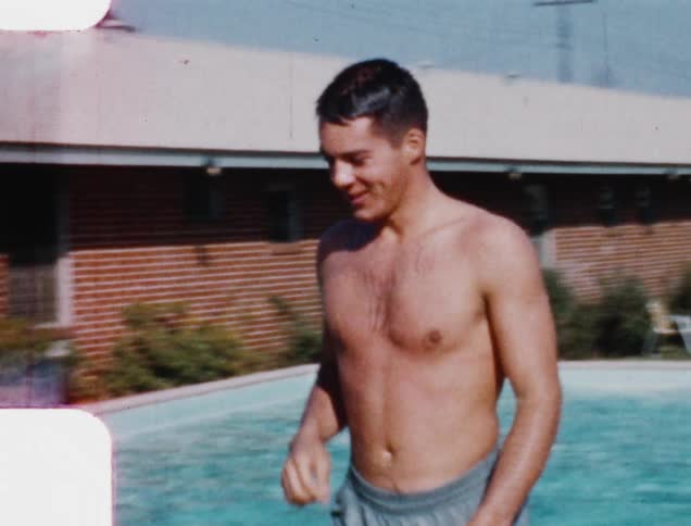 A young man in swimming trunks walks by a swimming pool in the backyard of a residence while smiling and waves at the camera