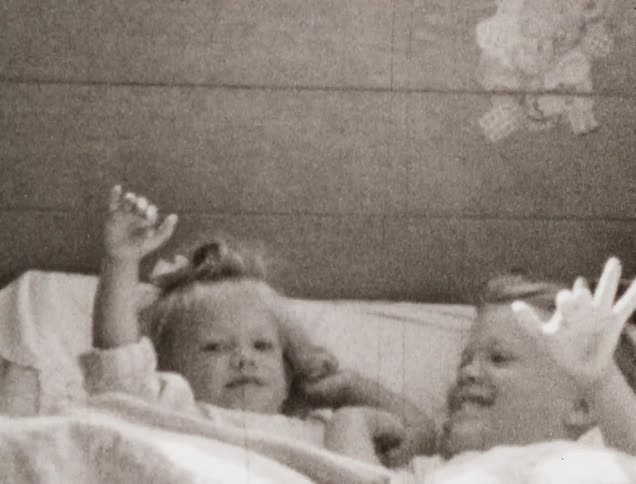 Two young girls lay on a bed and smile and wave