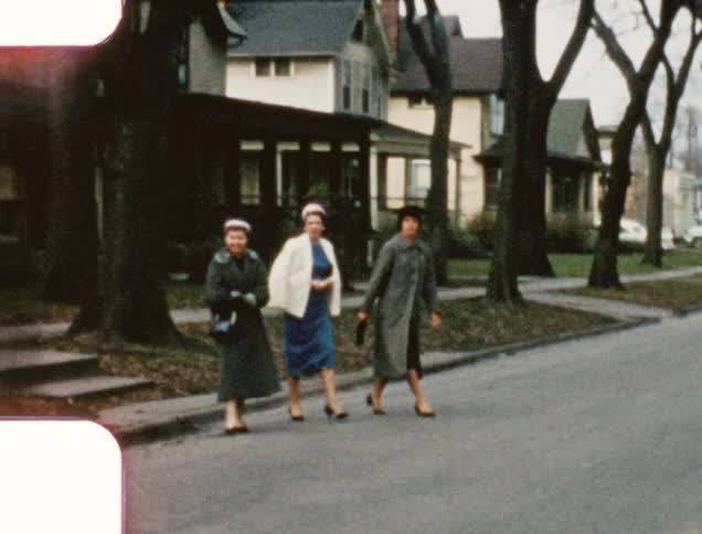 Three white woman in dresses and coats walk over to cars in a street