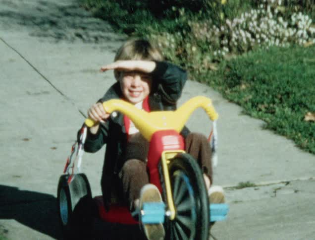 Boy riding a big wheel along the sidewalk of a neighborhood while using one hand and then the other to shield the sun from his eyes